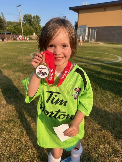 Ella wears a soccer jersey and holds up a medal from around her neck.