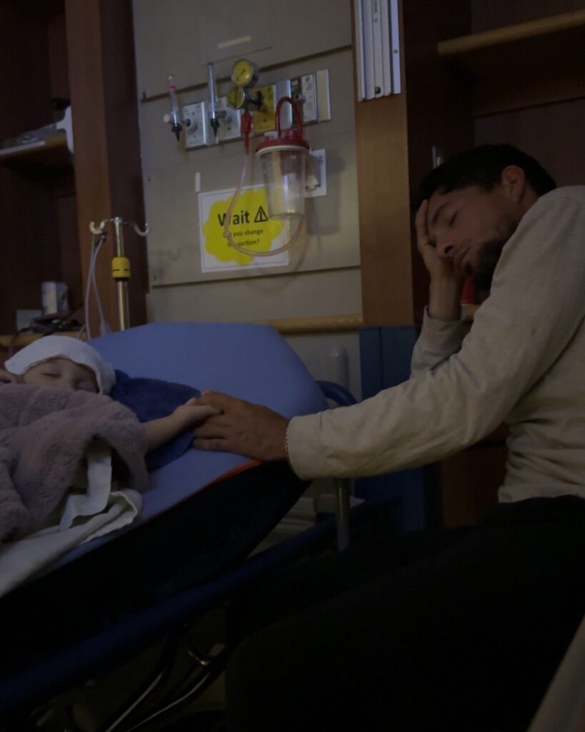 Ella is asleep in a hospital bed holding her dads hand who is asleep in a chair beside her bed.