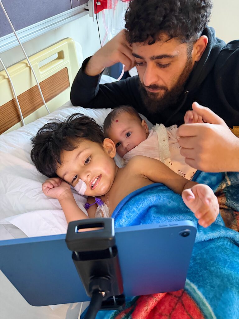 Rayan watches his tablet in his hospital bed next to his baby sibling and dad