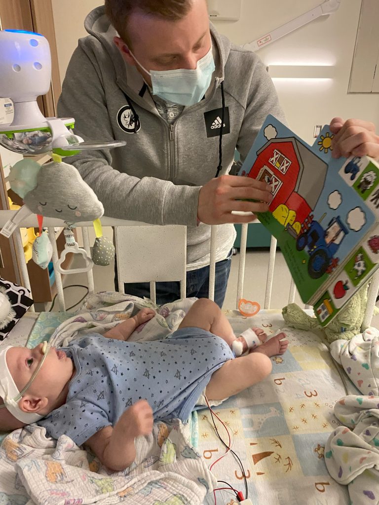 Paxton lies in his hospital crib while his Dad Mark reads to him.